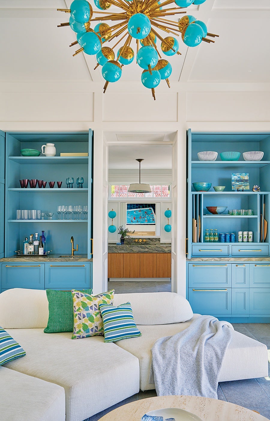 A modern living space featuring a white sectional sofa adorned with green, floral, and striped throw pillows, and a light gray blanket draped over one side. Behind the sofa are two built-in blue cabinets with gold handles, one containing glassware and bottles with a small sink, and the other holding bowls, cans, and bottles. A doorway between the cabinets leads to a bathroom with a wooden vanity and a large mirror reflecting a colorful painting. Above the living area hangs a striking chandelier with gold rods and turquoise spherical accents.