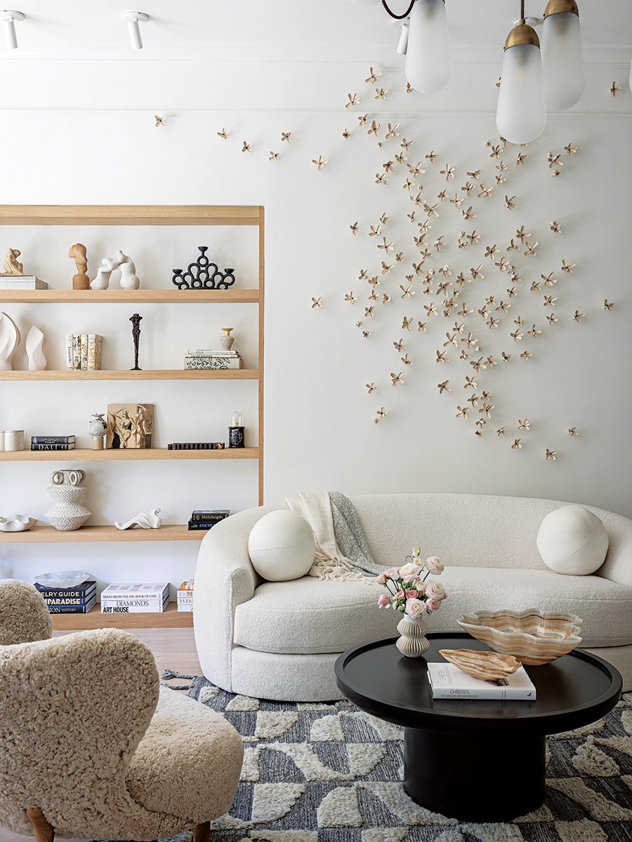 A modern living room features a curved white sofa with two round white pillows and a light throw blanket. In front of the sofa is a black round coffee table holding a small vase with pink flowers, two decorative shell-shaped bowls, and a book. A textured beige armchair is partially visible in the foreground. The wall behind the sofa is decorated with a cluster of small, gold, flower-like wall sculptures. To the left, a wooden open shelving unit displays various abstract sculptures, books, and decorative objects. The floor has a patterned rug in shades of gray and white. The ceiling has white track lights and hanging pendant lights with frosted glass shades and brass accents.