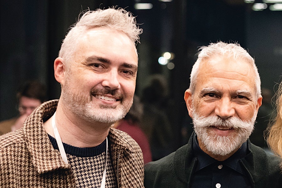 Two men with gray hair and beards are smiling. The man on the left has short, curly hair and is wearing a brown houndstooth jacket over a patterned sweater. The man on the right has a neatly groomed mustache and beard and is dressed in a dark jacket over a black shirt. The background is dimly lit with blurred figures and lights.
