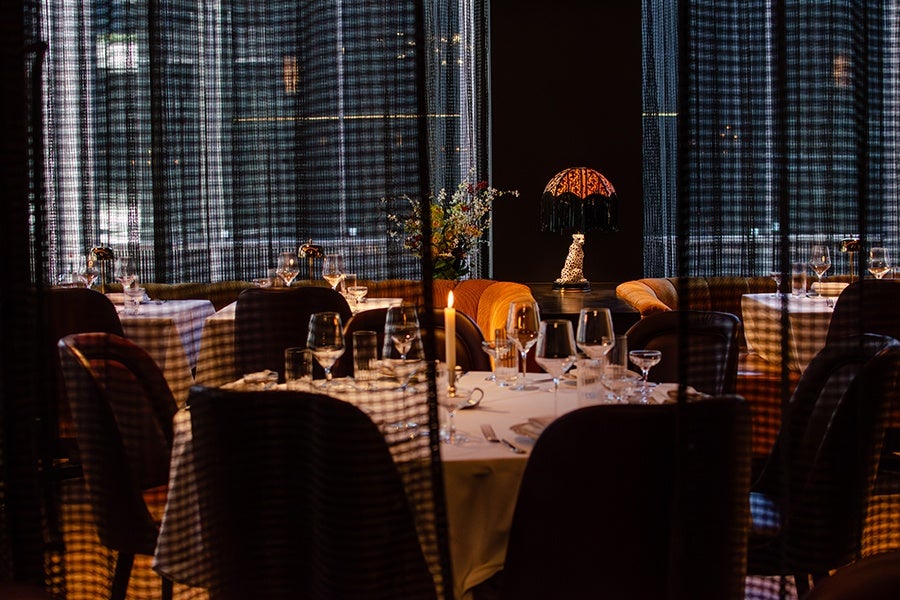 A dimly lit restaurant interior with round tables covered in white tablecloths, set with wine glasses, water glasses, and cutlery. A single lit candle is on the central table. Dark chairs surround the tables, and a decorative lamp with a fringed shade and a leopard figurine is visible in the background. Sheer curtains with a grid pattern partially obscure the view.