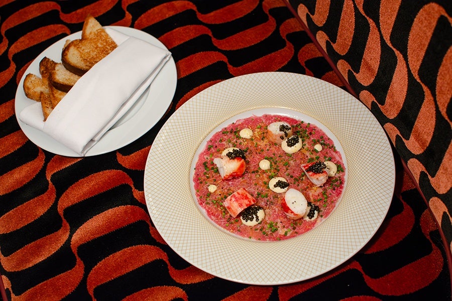 A white plate with a gold lattice pattern holds a dish featuring a pink base topped with pieces of seafood, dollops of cream, chopped chives, and black caviar. Next to it, a smaller white plate contains several slices of toasted bread partially wrapped in a white napkin. Both plates are placed on a fabric with a bold orange and black wavy pattern.