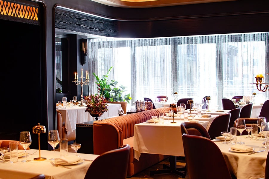 A stylish restaurant interior featuring tables covered with white tablecloths, set with wine glasses, plates, and napkins. The seating includes plush, brown upholstered chairs and a long, tufted brown banquette. Decorative elements include tall candelabras with lit candles and a floral arrangement. Large windows with sheer curtains allow natural light to fill the space.