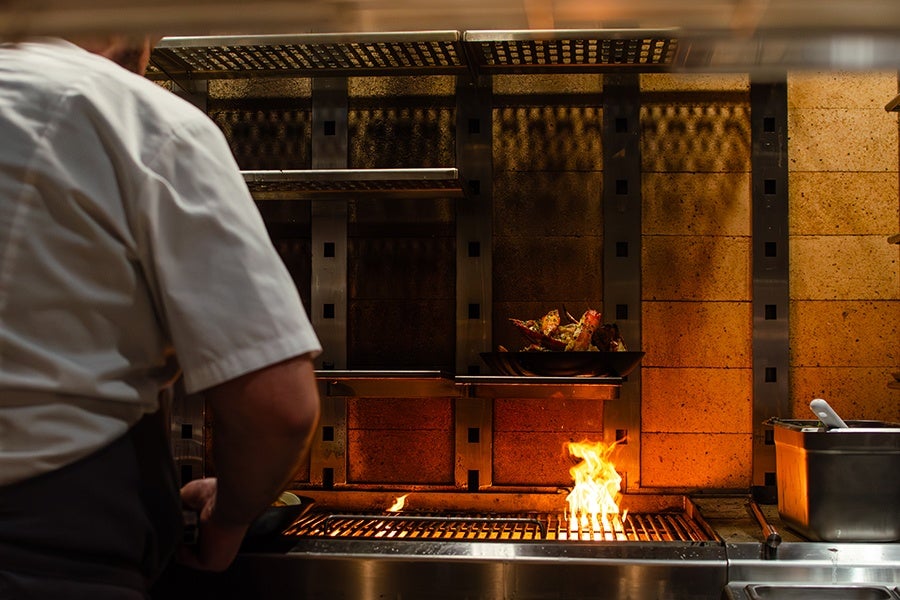 A chef in a white shirt and dark apron stands in front of a grill with an open flame. On a shelf above the grill, there is a black bowl containing cooked crab legs. The background features a tiled wall with metal supports and shelves. A metal container with a white utensil is positioned to the right of the grill.