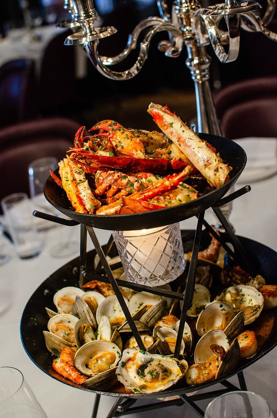 A two-tiered seafood platter featuring cooked crab legs and claws on the top tier, garnished with herbs. The bottom tier contains a variety of shellfish, including clams, shrimp, and scallops, all cooked and garnished with herbs. A lit candle in a glass holder is positioned between the two tiers, providing warm light. The platter is set on a white tablecloth with blurred background elements of a dining setting.