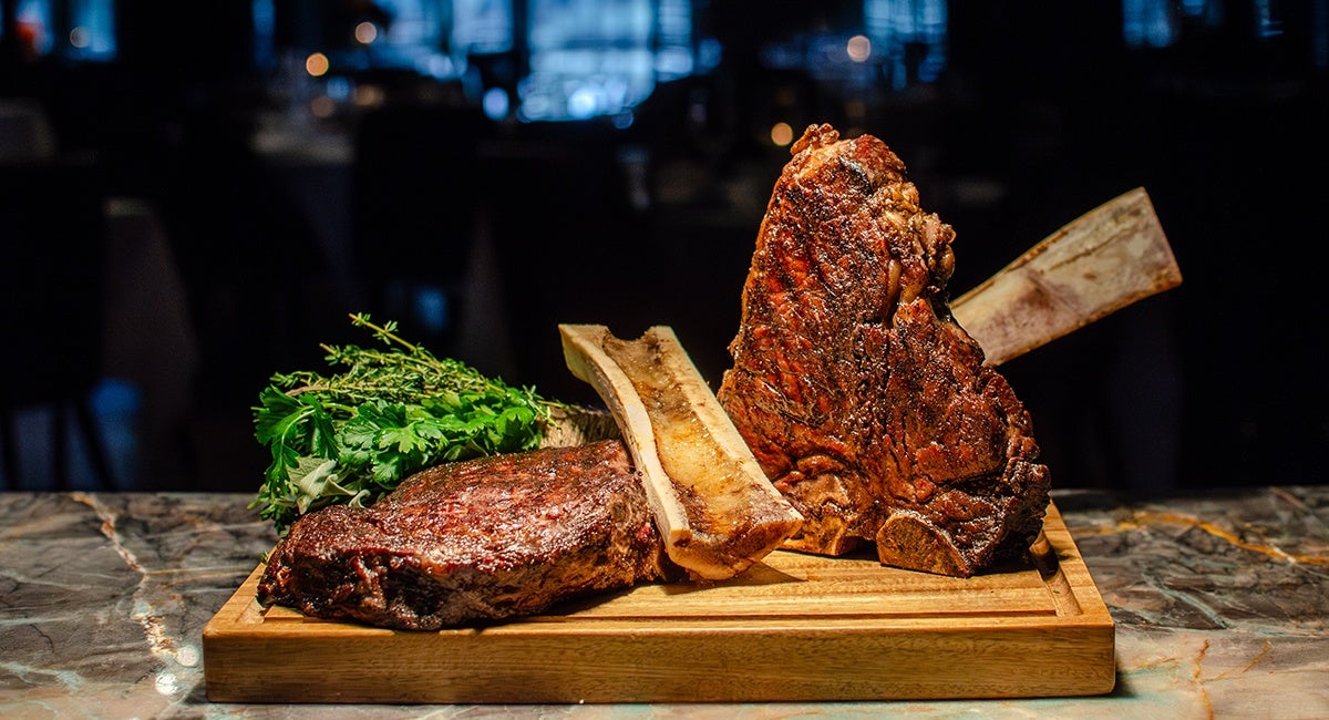 Two large grilled steaks with bones, placed on a wooden cutting board, accompanied by fresh green herbs on a marble surface with a dark, blurred background.