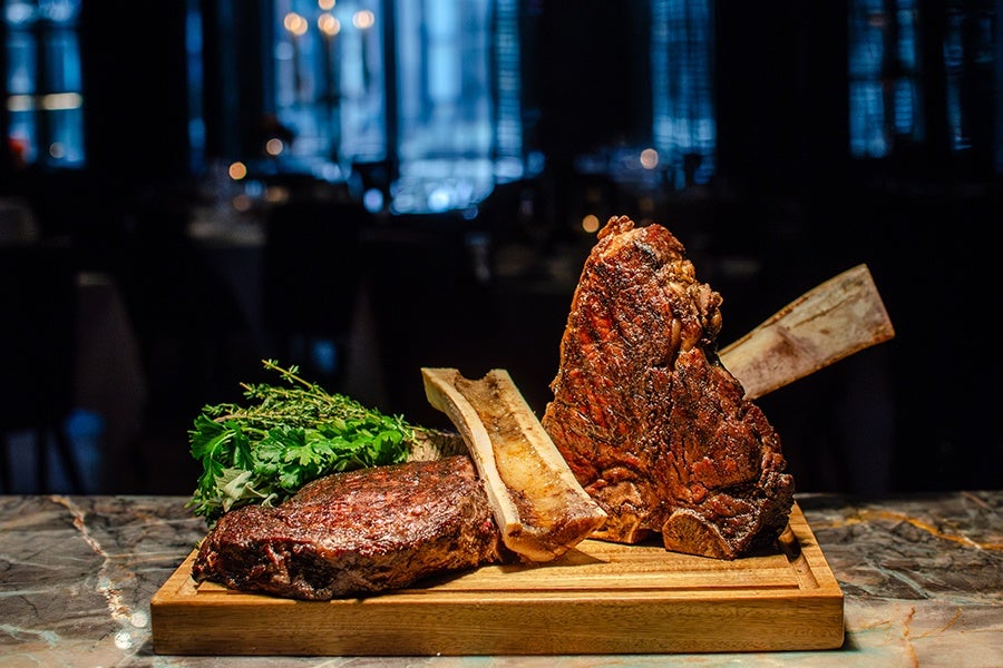 Two large, grilled bone-in steaks are presented on a wooden cutting board, accompanied by fresh green herbs. The steaks have a rich, charred crust and are positioned upright, showcasing the bones. The background is dark and blurred, highlighting the meat as the focal point.