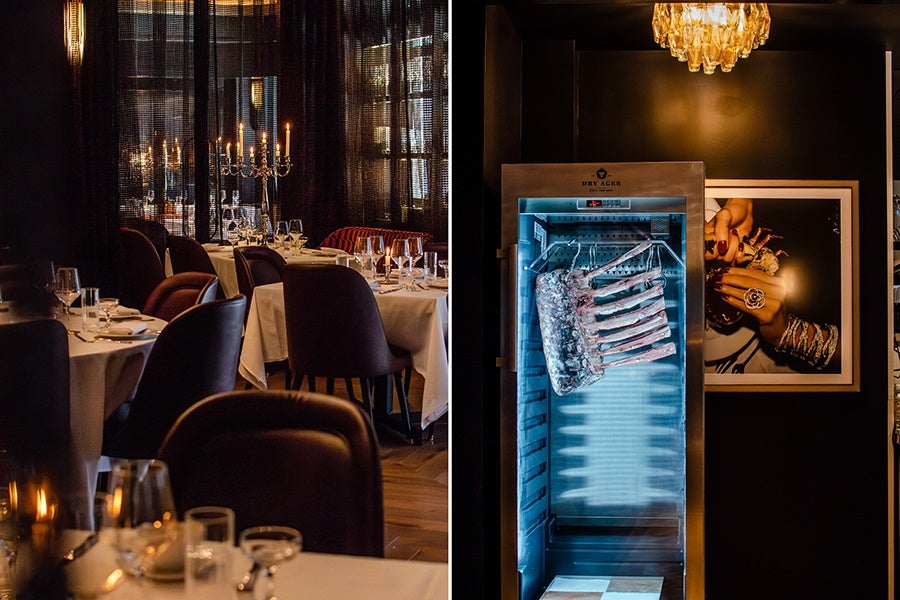 A dimly lit, elegant restaurant dining area with tables covered in white tablecloths, set with glassware and plates, and a candelabra centerpiece. Adjacent to this is a modern meat aging cabinet with a rack of ribs inside, illuminated with blue light, against a dark wall featuring a framed photograph of a hand adorned with rings and bracelets.