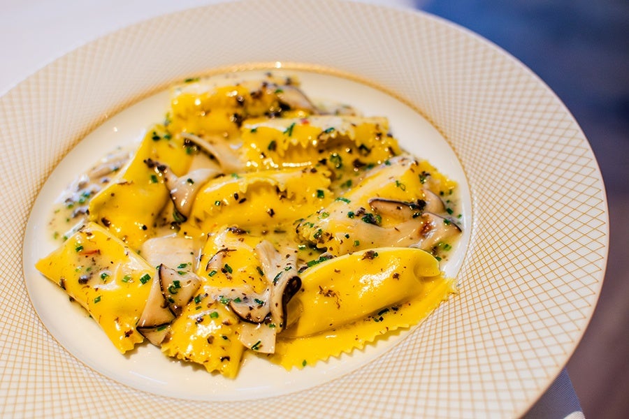Yellow ravioli pasta served with a creamy sauce, garnished with sliced mushrooms and finely chopped herbs, presented on a white plate with a gold geometric pattern around the rim.
