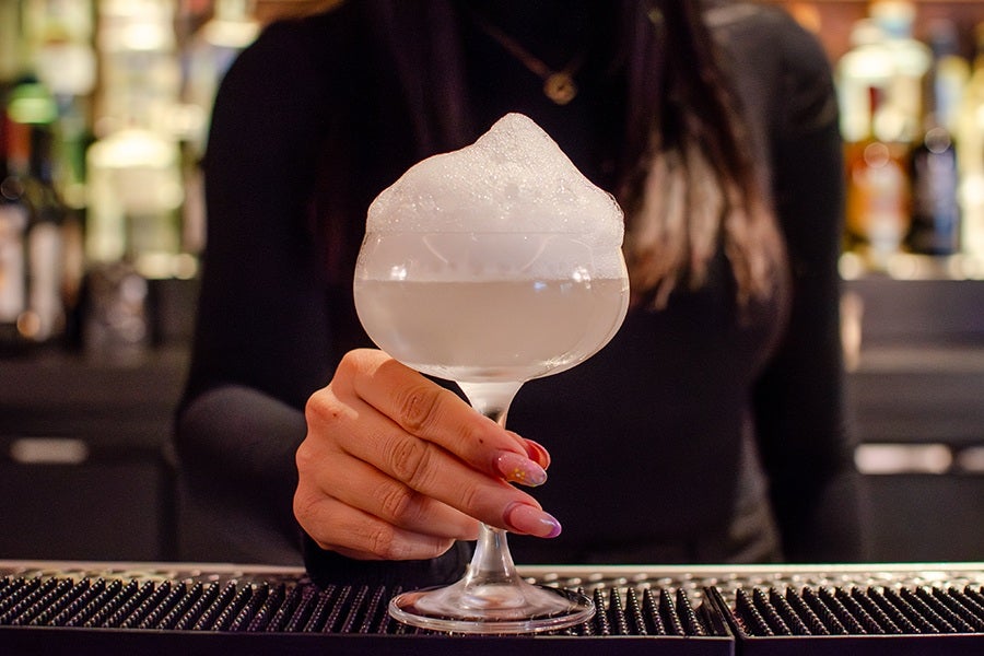 A hand with manicured nails holding a cocktail glass filled with a frothy white drink, set against a blurred background of a bar. The person is wearing a black long-sleeve top.