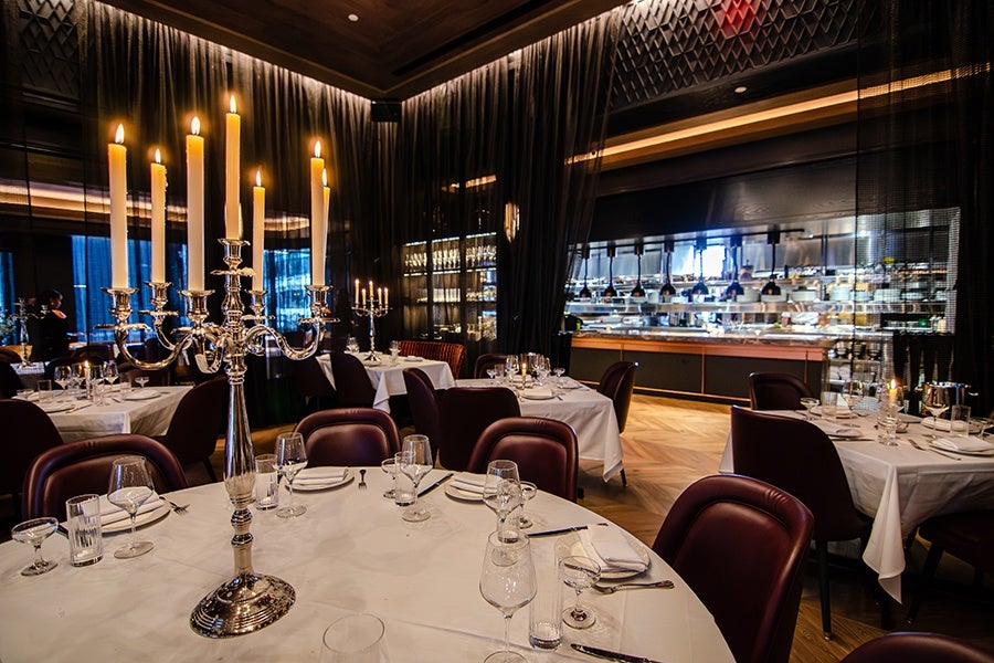 A fine dining restaurant interior with round tables covered in white tablecloths, set with glassware, plates, and silverware. Each table features a tall silver candelabra with lit candles. The seating consists of dark leather chairs. The background includes a modern open kitchen with stainless steel appliances and shelves, separated by sheer black curtains. The ambiance is warm and elegant with soft lighting.