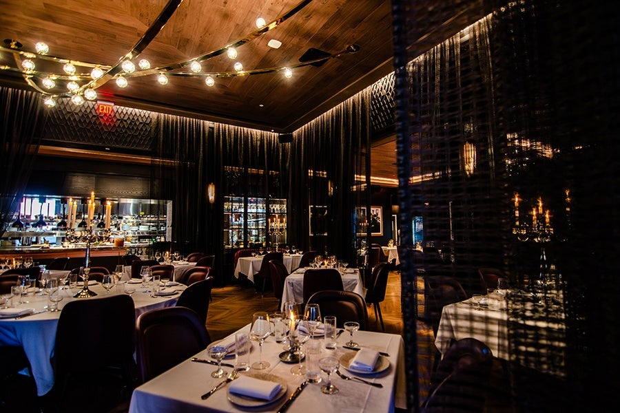 Dimly lit upscale restaurant interior with round and square tables covered in white tablecloths, set with glassware, silverware, and napkins. Dark curtains and wooden ceiling with string lights create a cozy atmosphere. A candelabra with lit candles is visible on one table, and a bar area can be seen in the background.