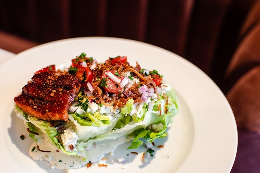 Wedge salad featuring a quarter head of iceberg lettuce topped with crumbled blue cheese, chopped cherry tomatoes, diced red onions, crispy fried onions, and chopped herbs, served with a creamy dressing on a white plate.