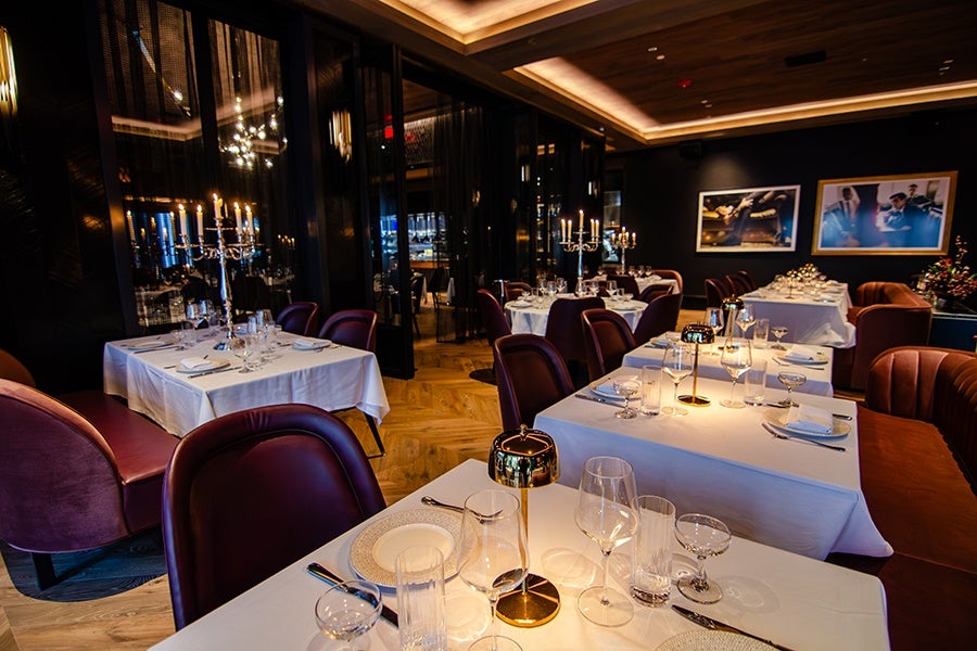 A dimly lit, elegant restaurant interior with tables covered in white tablecloths. Each table is set with glassware, plates, cutlery, and small gold lamps. Some tables have candelabras with lit candles. The seating includes plush, dark red and brown chairs and booths. The walls are dark, adorned with framed artwork, and the ceiling features recessed lighting. The floor is wooden with a herringbone pattern.