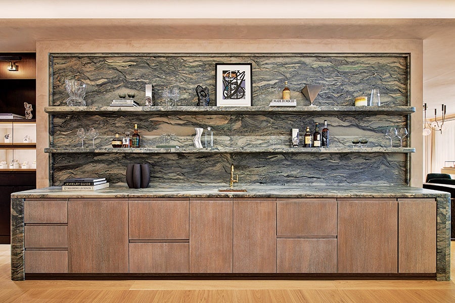 A modern bar setup features a long wooden cabinet with a green and gray marble countertop and matching marble backsplash and shelves. The two marble shelves hold various decorative items, including glassware, bottles of liquor, books, and art pieces. A small gold faucet is centered on the countertop. The overall design combines natural stone textures with sleek wood cabinetry.