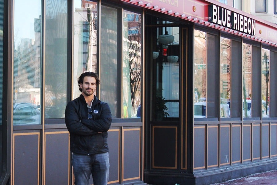 A man with dark hair and a beard stands with his arms crossed in front of a building with large windows and a sign that reads "BLUE RIBBON." He is wearing a black jacket and jeans. The building exterior features dark panels with gold trim.