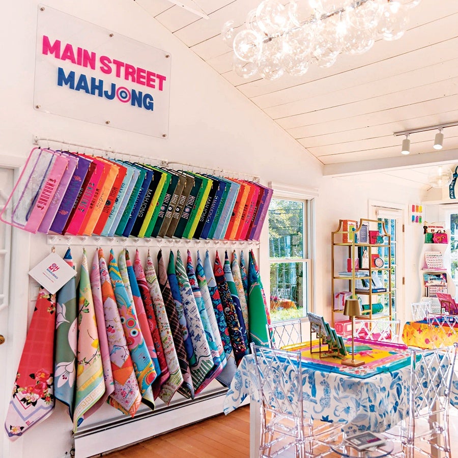 Bright, colorful scarves and pouches are displayed on racks in a well-lit, modern store with a sign reading "Main Street Mahjong." The space features clear acrylic chairs around tables covered with patterned cloths, and shelves with various vibrant items. Large windows let in natural light, highlighting the cheerful and inviting atmosphere.