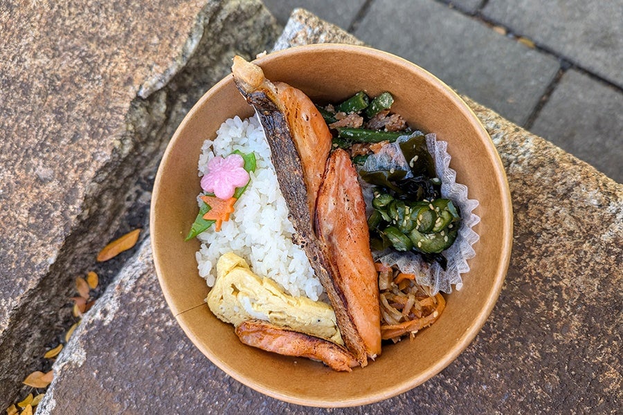 A brown bowl containing a meal with grilled salmon fillet, white rice topped with pink and orange flower-shaped garnishes, a rolled omelette, sautéed green beans with ground meat, seaweed salad with cucumber slices, and a portion of stir-fried vegetables. The bowl is placed on a rough stone surface outdoors.