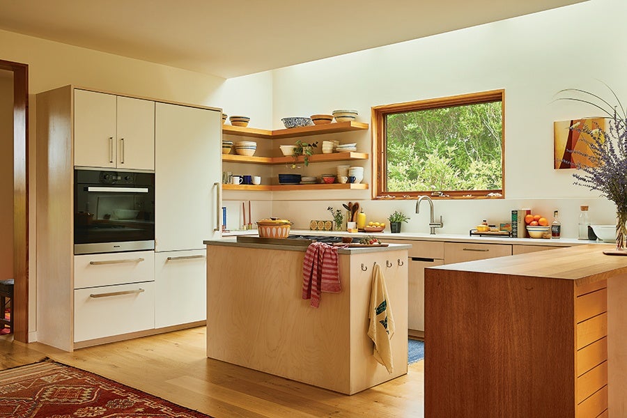 Modern kitchen with light wood cabinetry and a central island featuring a cooktop and hanging towels. Open wooden shelves hold various bowls and cups. A large window above the sink offers a view of green foliage. The floor is light wood, and a patterned rug is partially visible. A wooden countertop extends from the right side, and a vase with purple flowers sits on it.