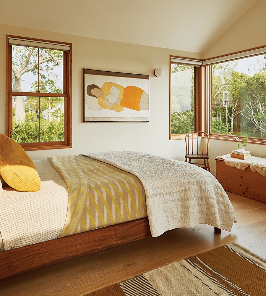 Bedroom with a wooden bed frame featuring beige and yellow bedding, a mustard yellow pillow, and a cream quilt. The room has large windows with wooden frames, a wooden chair, a wooden chest with a sheepskin cover, and a painting of a reclining figure in orange and beige tones on the wall. The floor is light wood with a patterned rug in shades of brown and beige.