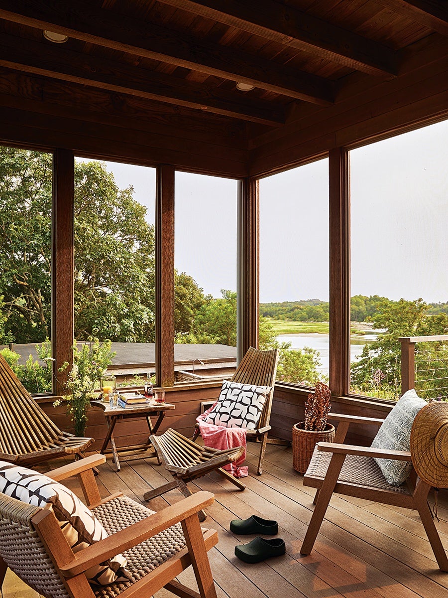 Wooden screened porch with four wooden chairs, each with patterned cushions, and a small wooden table holding drinks and books. A pair of black clogs is on the wooden floor, and a woven basket with dried plants is in the corner. The porch overlooks a lush green landscape with trees and a body of water in the distance.