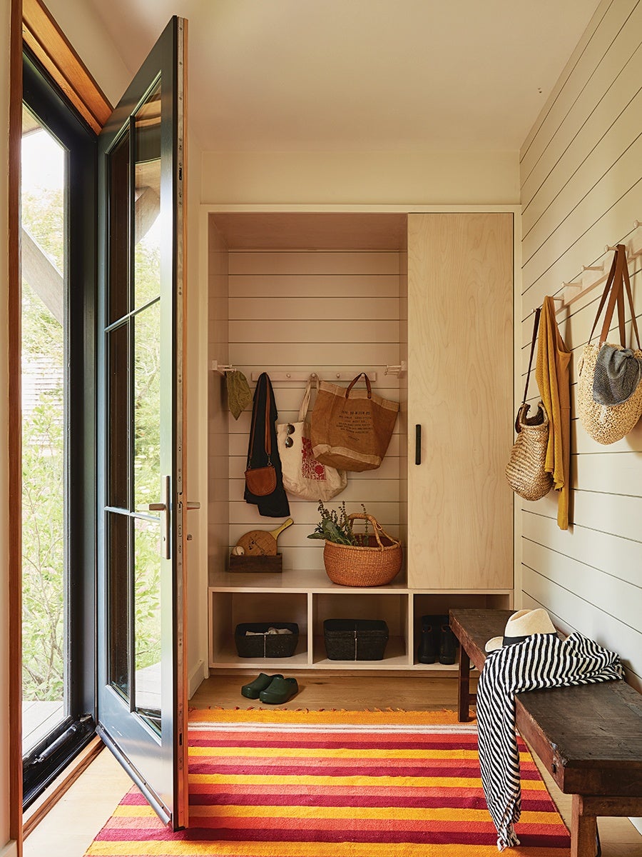 Entryway with a wooden bench draped with a black and white striped cloth and a white hat. A colorful striped rug in shades of red, orange, and yellow covers the floor. A built-in wooden storage unit with cubbies and a cabinet holds baskets, boots, and hanging bags. Wall hooks on the right side hold woven bags and a mustard yellow scarf. The door on the left is open, showing a glimpse of greenery outside. The walls are light-colored with horizontal paneling.