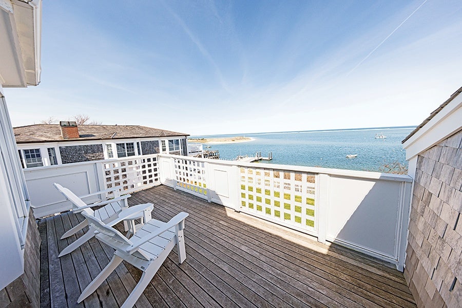 Wooden deck with two white Adirondack chairs overlooking a calm blue ocean, with a neighboring building and a small dock visible in the background under a clear sky.