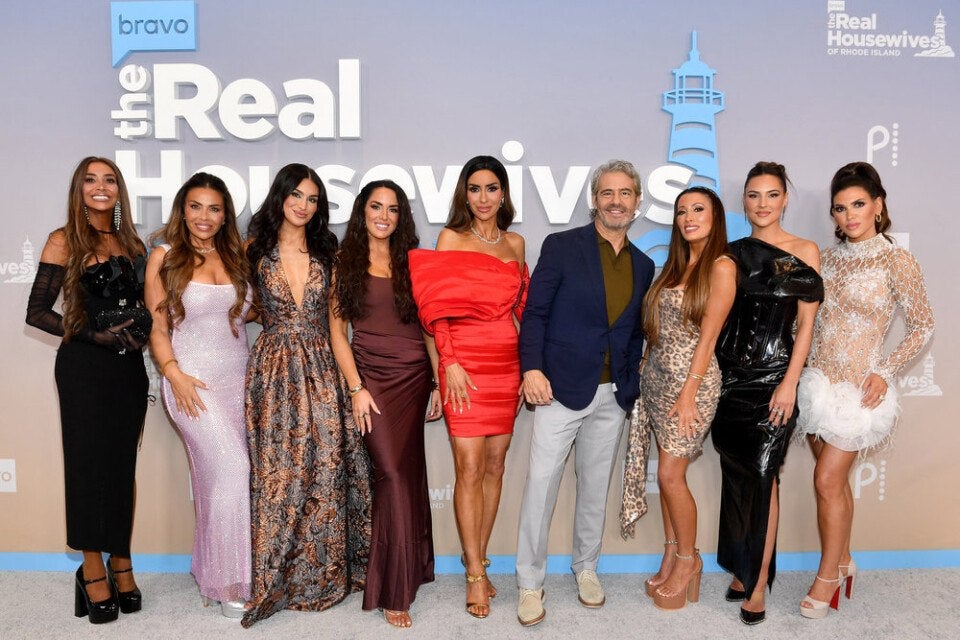 A group of nine people posing in front of a backdrop with "The Real Housewives" logo. The group includes eight women and one man. The women are dressed in a variety of elegant outfits: one in a black dress with sheer sleeves, one in a light pink sparkly gown, one in a patterned sleeveless dress, one in a dark maroon dress, one in a bright red off-shoulder dress, one in a leopard print dress, one in a shiny black dress, and one in a white lace dress with feather details. The man is wearing a navy blazer, olive green shirt, and light gray pants. They are all standing on a light-colored floor with a blue border at the bottom of the backdrop.