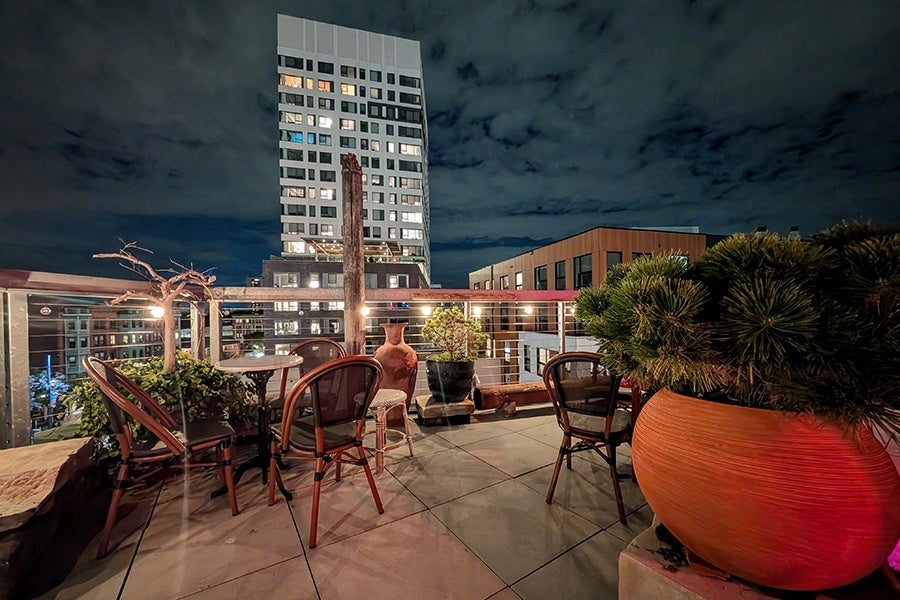 Rooftop patio at night featuring a tiled floor, several chairs, and a small round table. The patio is decorated with various potted plants, including a large orange pot with dense greenery on the right. In the background, a tall modern building with illuminated windows stands against a cloudy night sky.