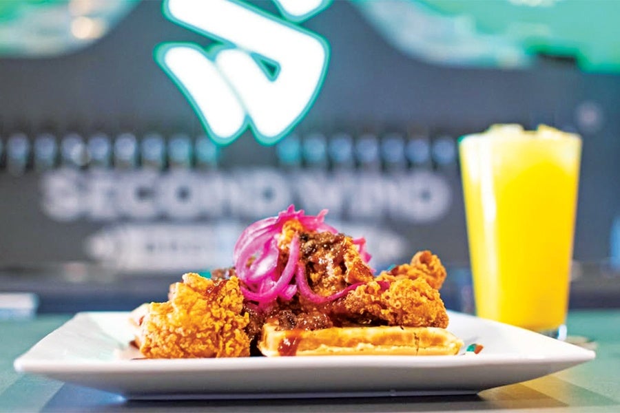 Crispy fried chicken pieces served on a waffle, topped with pickled red onions and sauce, on a white rectangular plate. A glass of orange juice with ice is placed in the background.