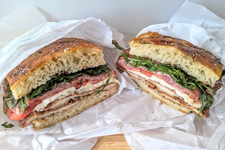 Two halves of a sandwich on focaccia bread, filled with chicken, fresh tomato slices, leafy greens, prosciutto, and mozzarella cheese, wrapped in white parchment paper.