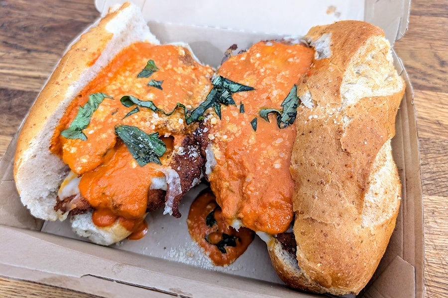Two breaded chicken sandwiches topped with orange sauce, grated cheese, and chopped fresh basil leaves, served in a cardboard takeout container.