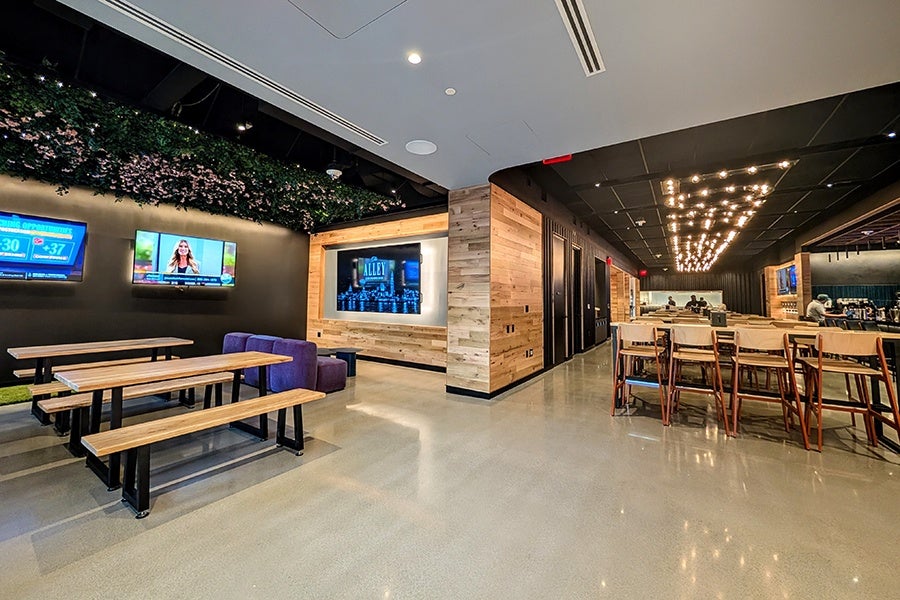 Modern lounge area with polished concrete floors, wooden picnic-style tables with benches, and a row of high chairs at a counter. The walls feature wood paneling and dark paint, with multiple flat-screen TVs mounted. Ceiling has black panels with string lights and a section decorated with greenery.