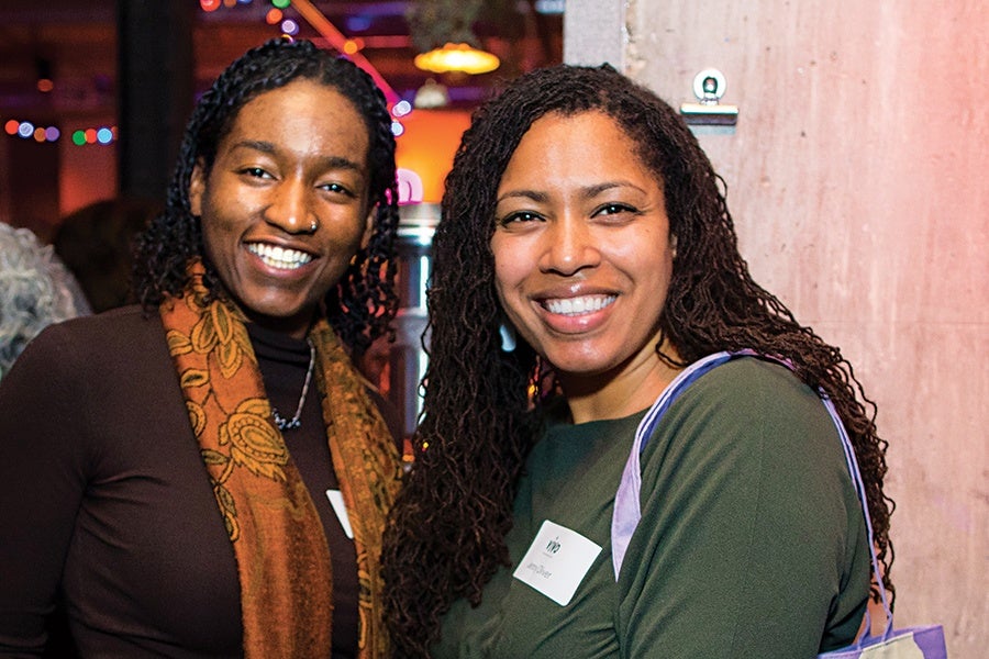 Two women smiling and posing together indoors. The woman on the left has dark curly hair, is wearing a dark brown top and an orange patterned scarf. The woman on the right has long curly hair, is wearing a green top, and has a name tag on her chest. The background is softly lit with colorful lights.