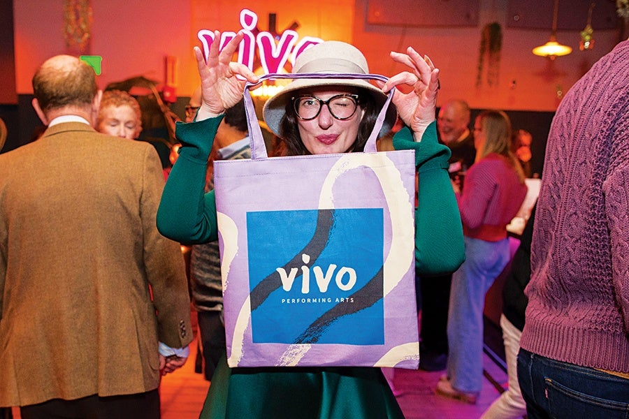 A person wearing glasses and a white hat is holding up a purple tote bag with a blue square and the words "vivo PERFORMING ARTS" printed on it. The person is winking and standing in a crowded indoor setting with warm lighting.