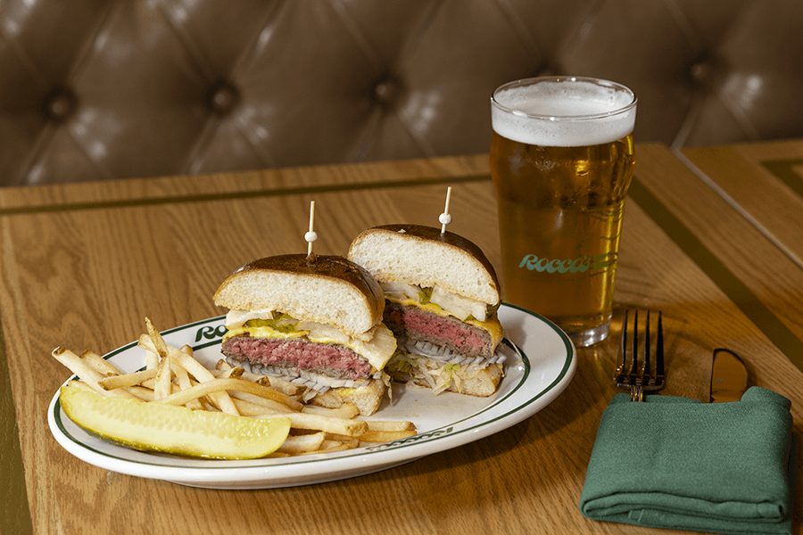 A plate with a cheeseburger cut in half, French fries, and a pickle spear. Next to the plate is a glass of beer with a foamy head. A green cloth napkin holds a fork and knife on the wooden table. The background features a tufted brown leather bench.